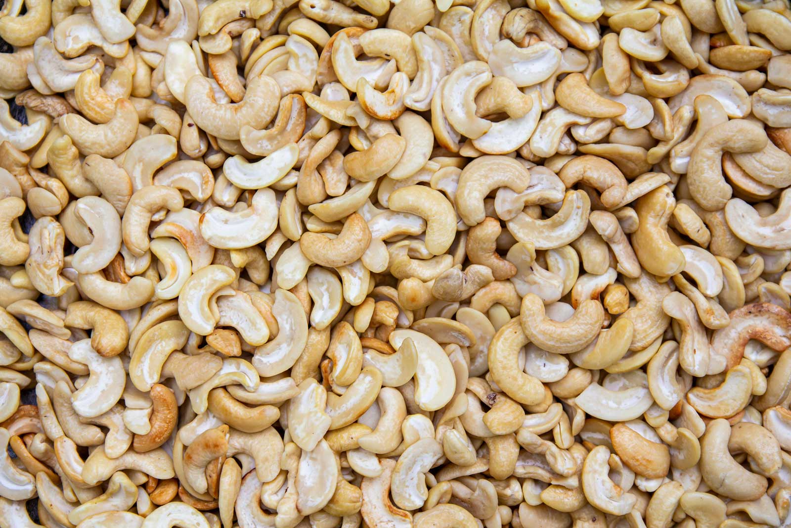 Raw cashew nuts spread out in a dense layer, showing their curved shape, pale golden color, and natural surface texture