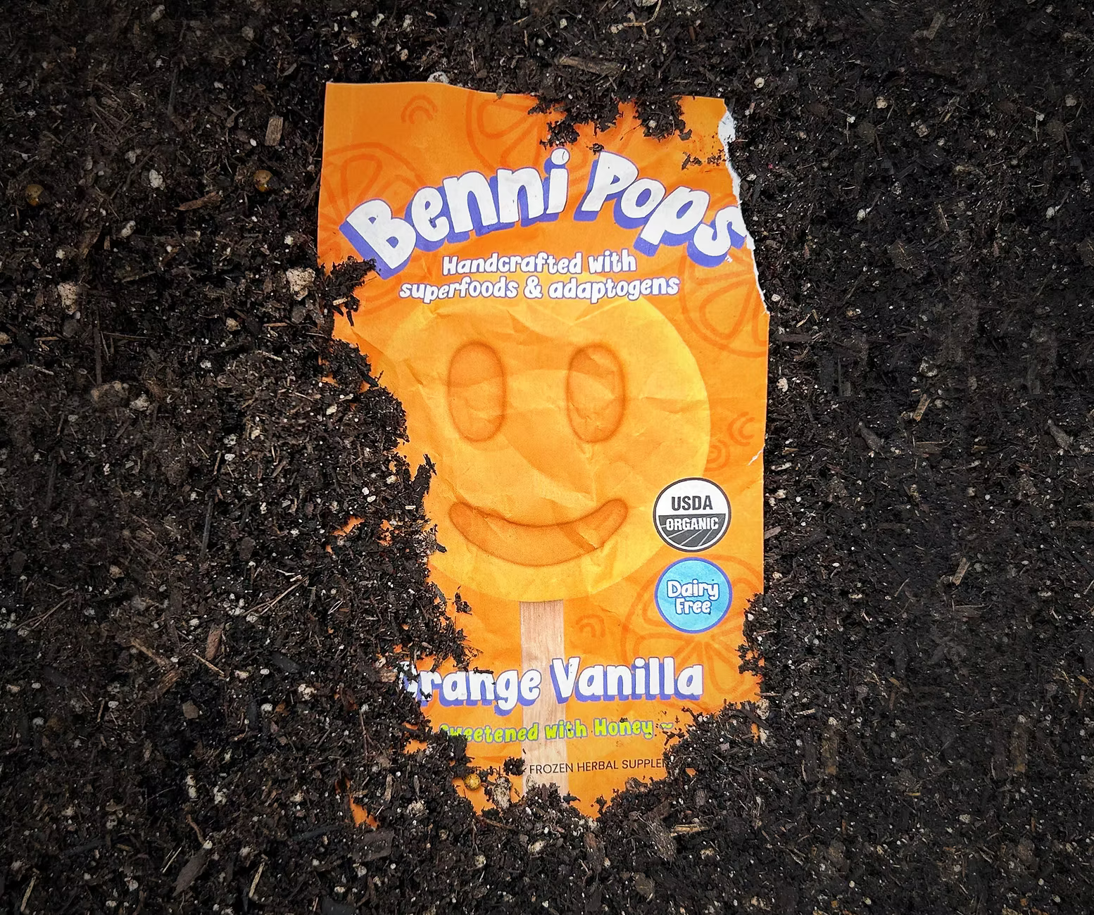 Benni Pops Orange Vanilla frozen dessert packaging placed on dark soil, highlighting organic, dairy-free ingredients and playful smiley-face design
