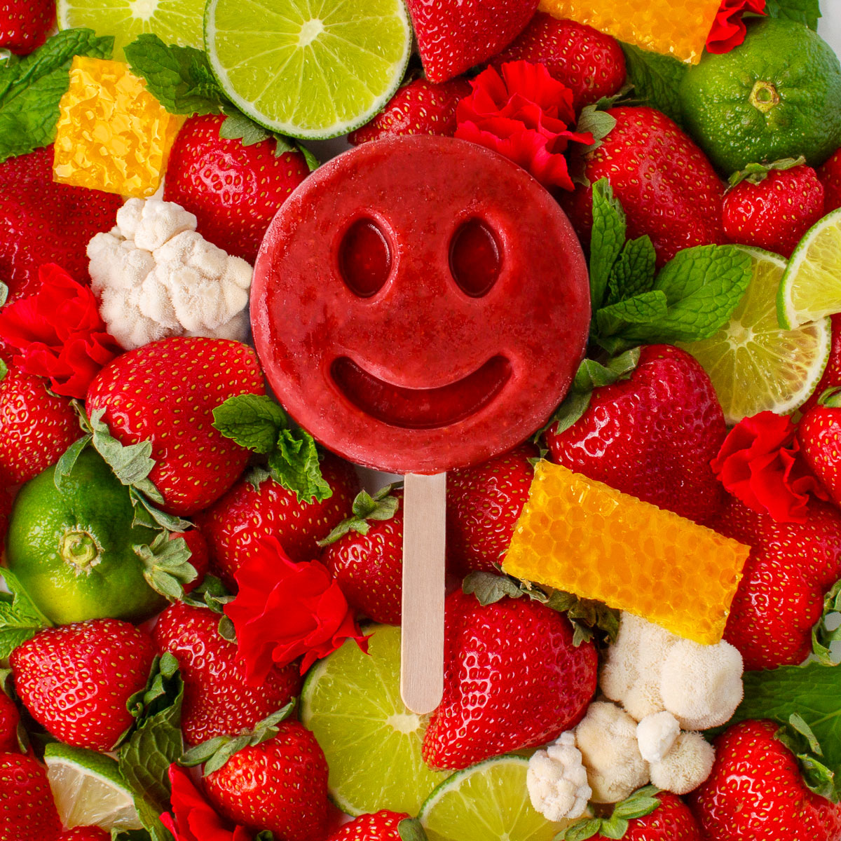 Red smiling frozen dessert on a stick surrounded by fresh strawberries, limes and honey