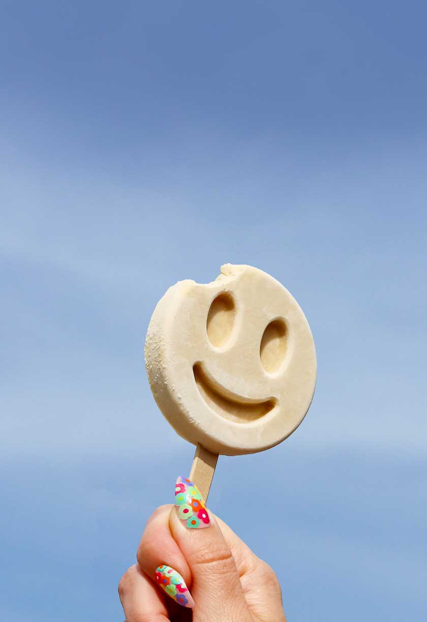 Hand holding a Benni Pops Orange Vanilla smiley face popsicle against a blue sky