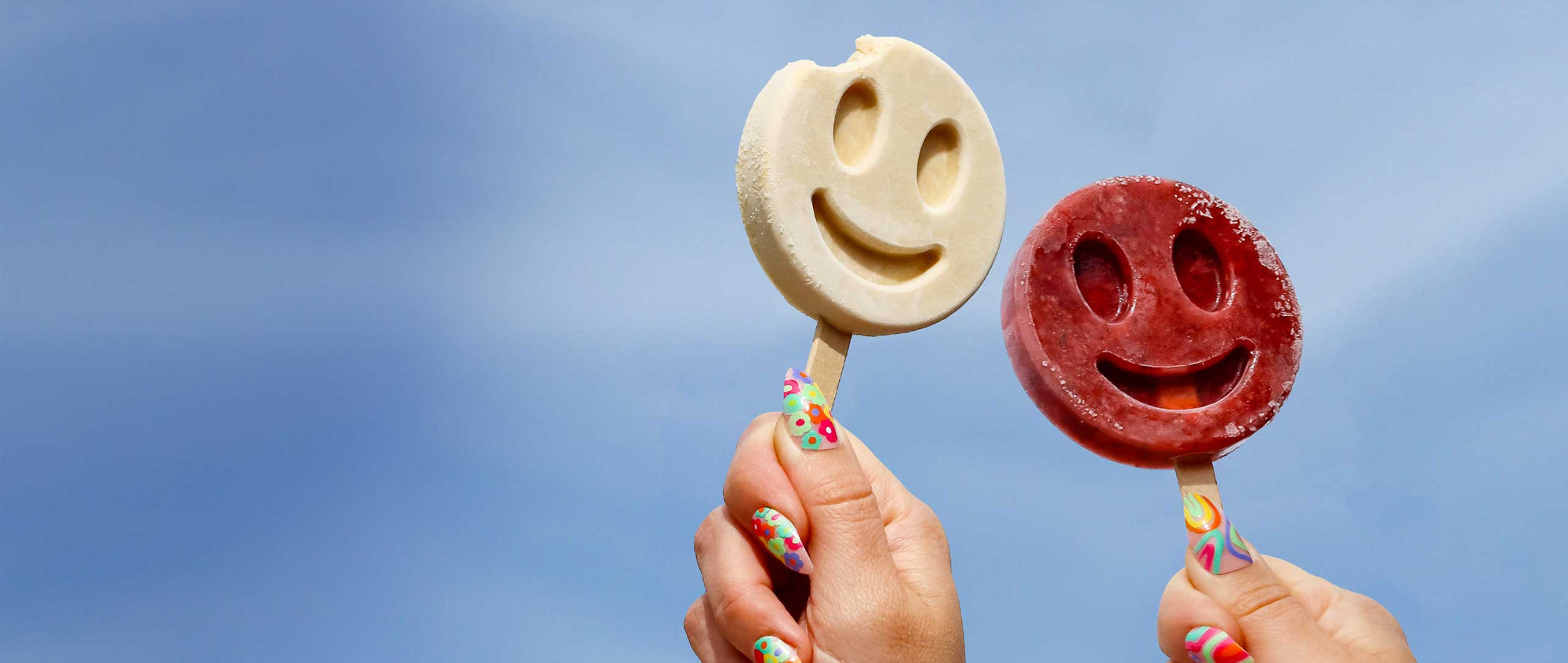 Two Benni Pops smiley face popsicles in Orange Vanilla and Strawberry Mint held against a blue sky