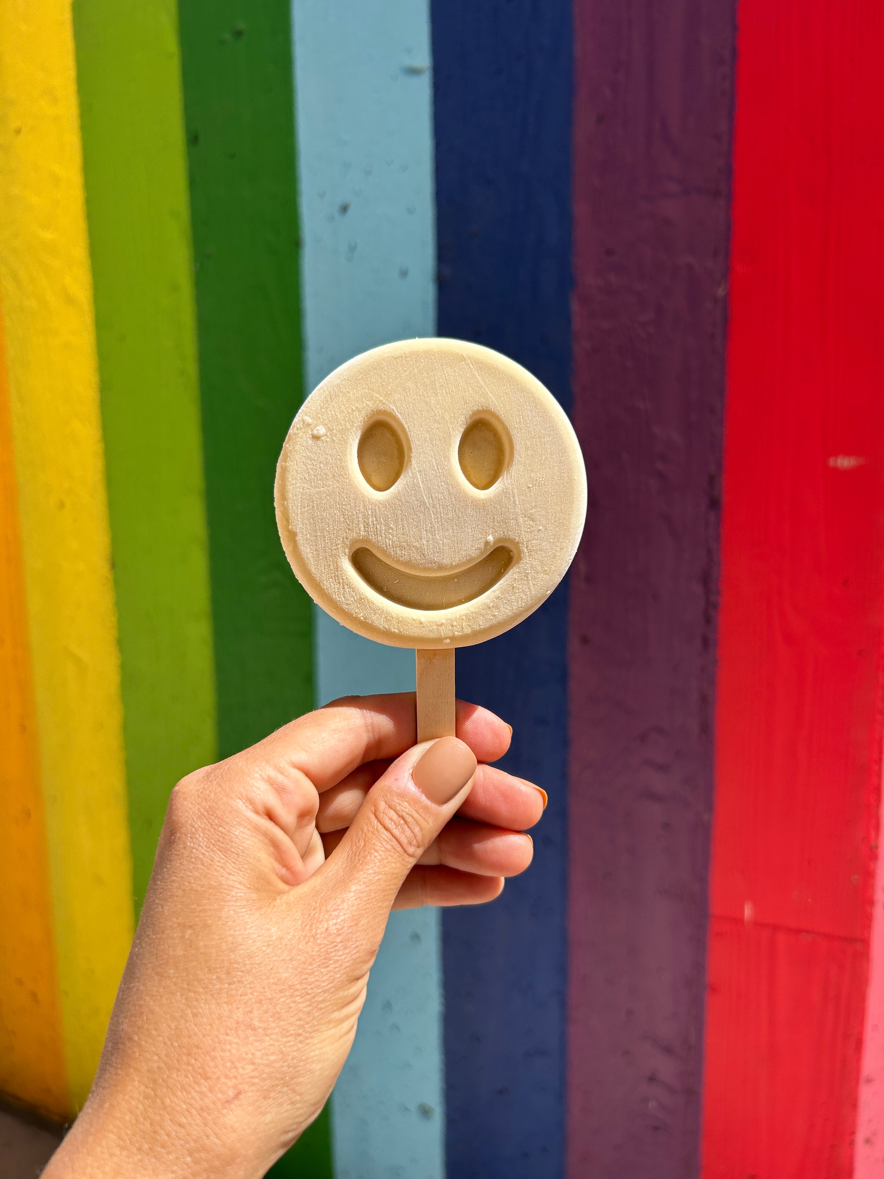 Hand holding a smiley-face frozen dessert in front of a rainbow-striped background