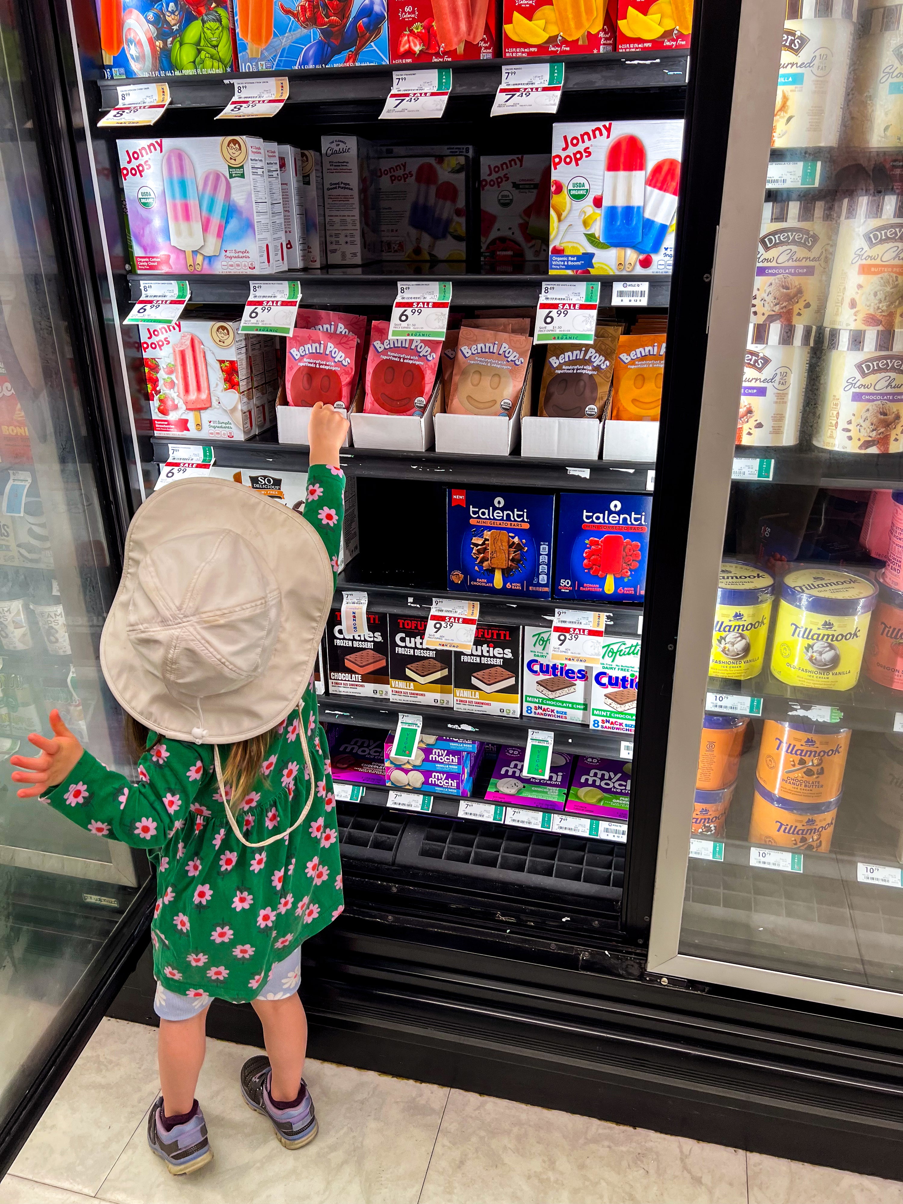Young child standing in front of a freezer case, looking at frozen desserts and grabbing a Benni Pop in a grocery store