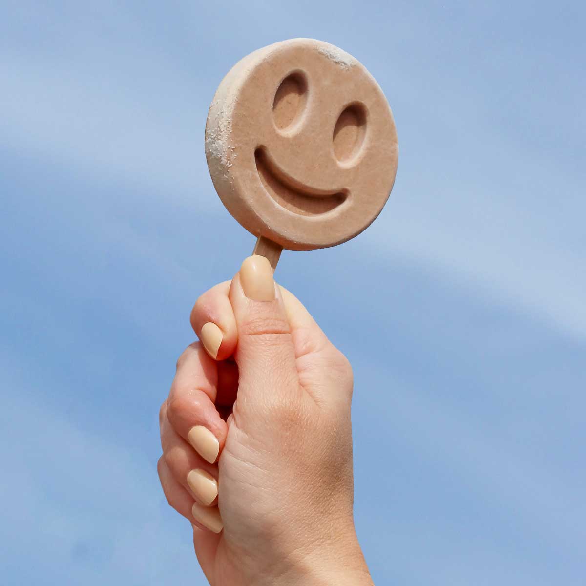 Hand holding a Benni Pops Cinnamon Roll smiley face popsicle against a clear blue sky