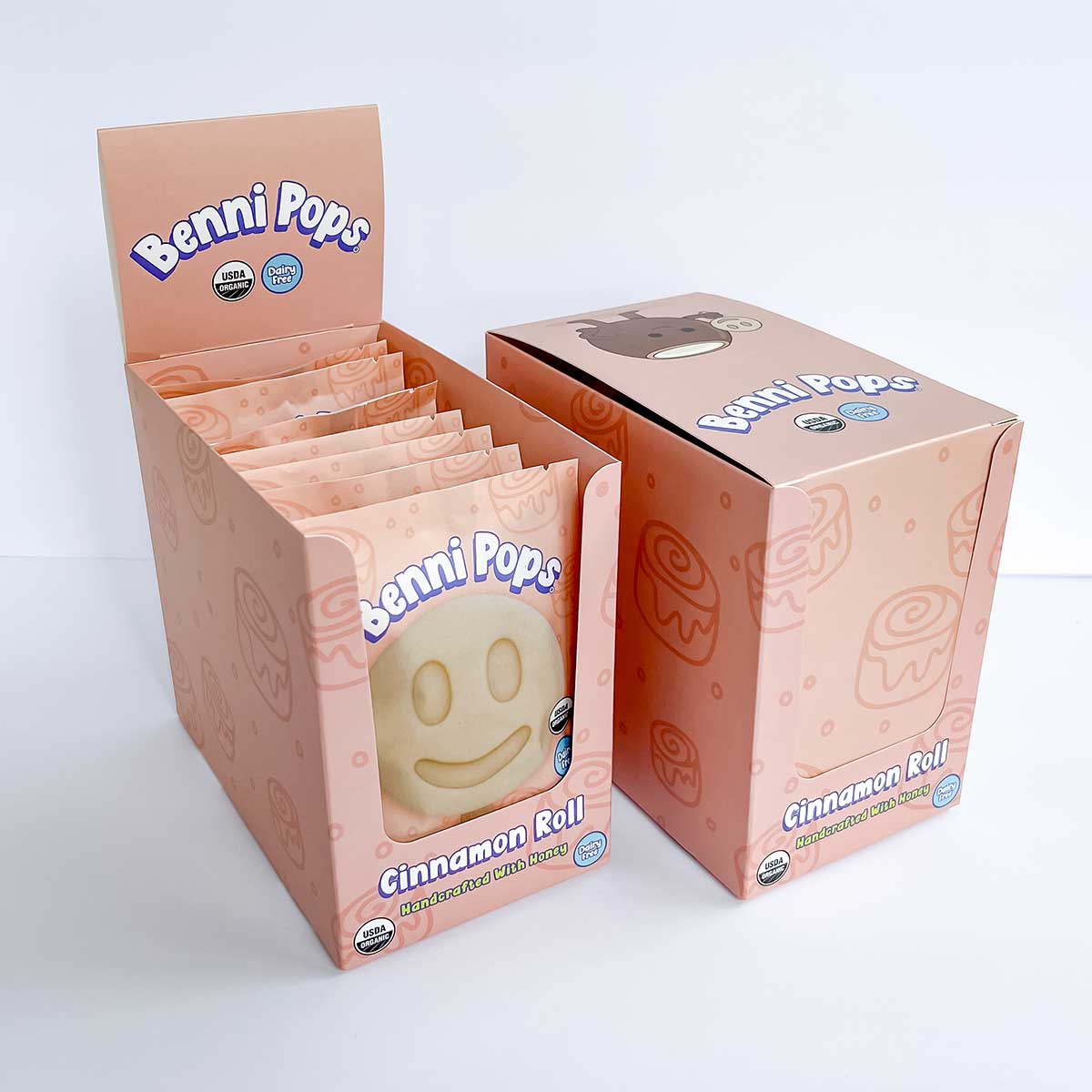 Benni Pops Cinnamon Roll is an organic, dairy-free frozen dessert made with honey and packaged in a playful branded retail display box. The image shows the Cinnamon Roll flavor in its individual wrapper alongside the matching Benni Pops case packaging for grocery freezer displays.
