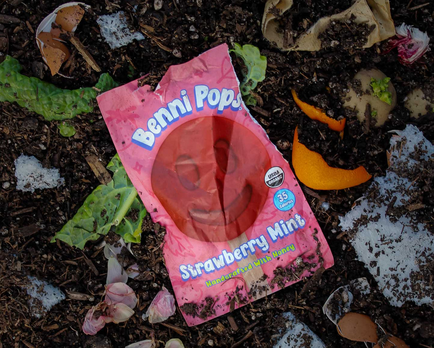 Benni Pops Strawberry Mint packaging partially buried in compost with food scraps and cruz foam pieces, highlighting compostable packaging