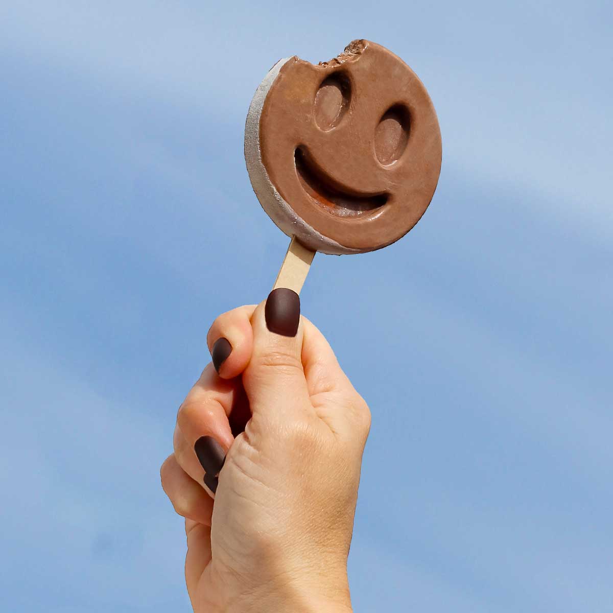Hand holding Benni Pops Fudge smiley faced popsicle against blue sky background