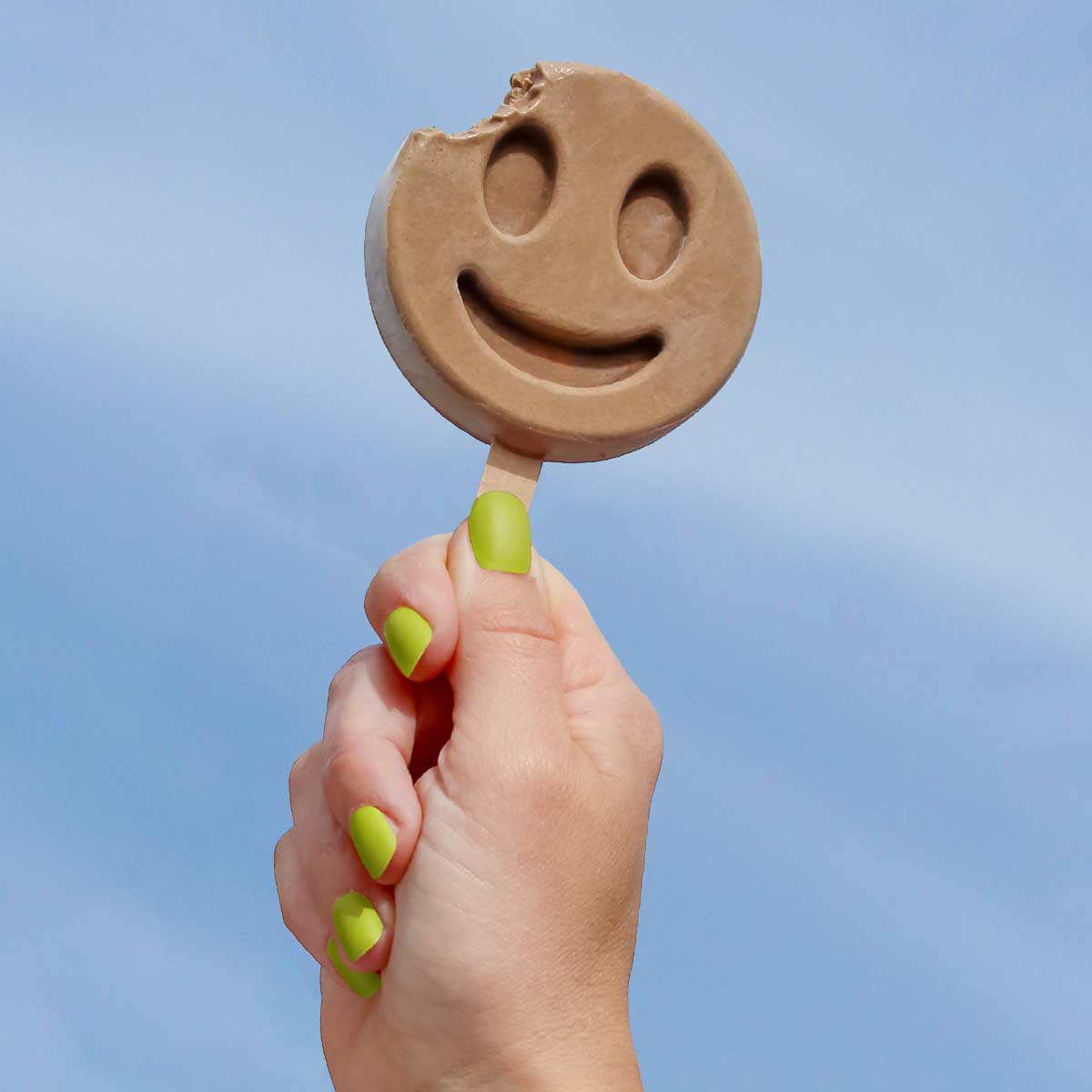 Hand holding a Benni Pops Latte popsicle with a smiley face against a clear blue sky