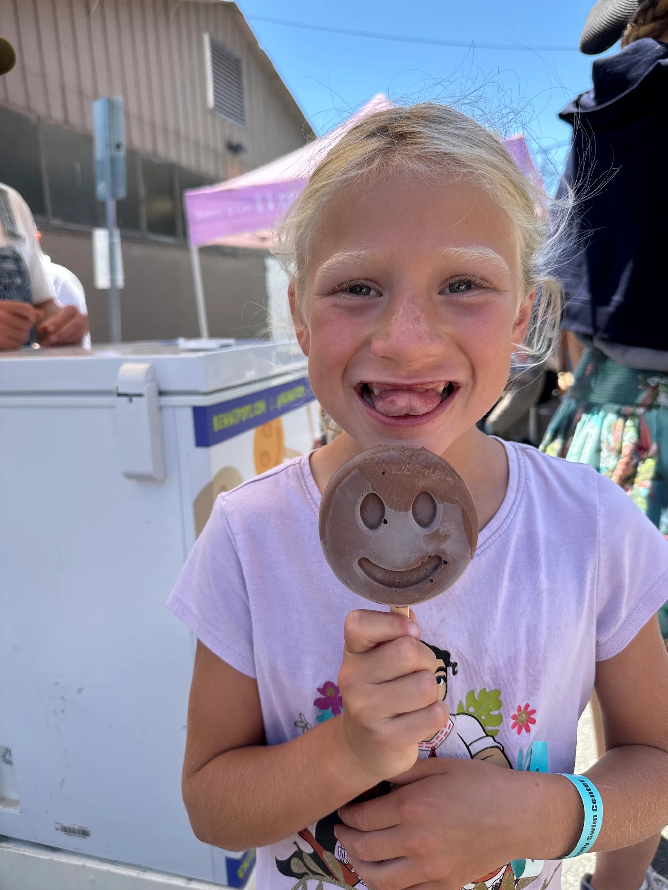 Child smiling and holding a smiley-face frozen dessert at an outdoor festival